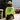 A construction worker viewed from behind, wearing a bright yellow hard hat, a neon yellow-green long-sleeve shirt with 'LONGVIEW BUILDERS' printed on the back, khaki pants, and work boots on a construction site.